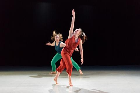 Two dancers, one in a rust jumpsuit with right arm extended straight up and another behind wearing shades of green with both arms outstretched