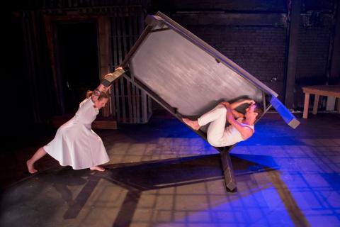 Dancer in a dress holds up one side of a sideways box containing a crouching dancer in pants.