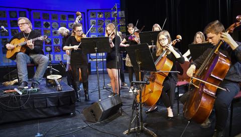 A large group of musicians of all ages play a variety of stringed instruments onstage in a black box theater.