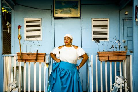 A woman, in a white head wrap and blue dress, stanfds before a doorway with flower boxes and carved lambs.