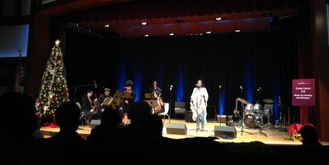 Image of a woman singing into a microphone from a stage, accompanied by musicians. A Christmas tree is on the left.