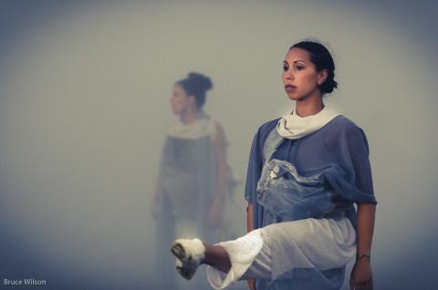 A dancer lifts her leg, staring straight ahead, while another dancer deeper in the fog is less visible