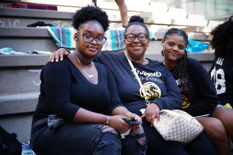 Three young Black women pose, while sitting on some stone steps outside.