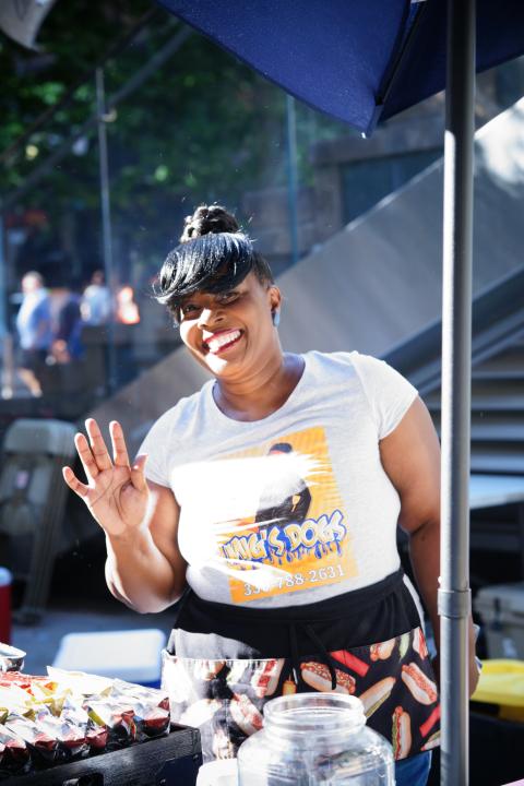 A Black woman with long bangs waves at the camera, while working behind a food stand.