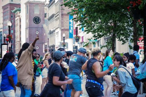 Outside, in a city center, a crowd dances.