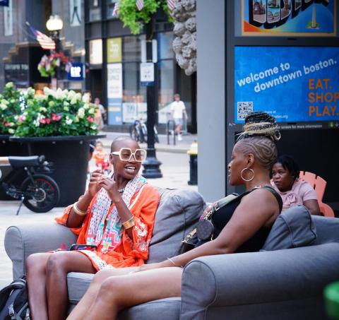 On a city street, two Black women sit on a couch together and chat.