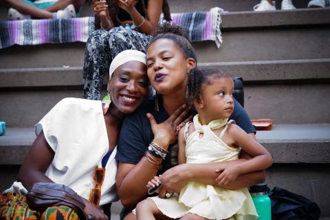 Two Black women poses with a Black toddler. One holds her chest and sings.