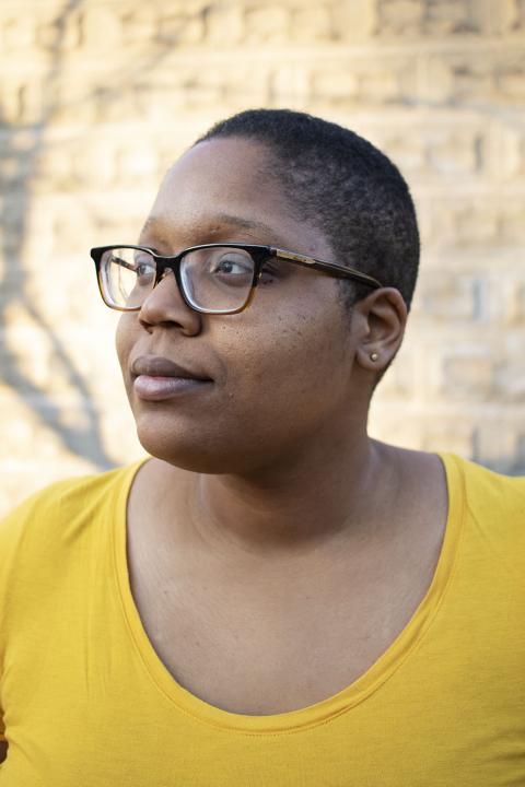 Deidra looks to the right and wears a yellow top. Deidra is Black, has short hair, and wears glasses.
