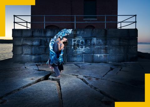 A white woman throws her dress back on top of a stone building next to the water.