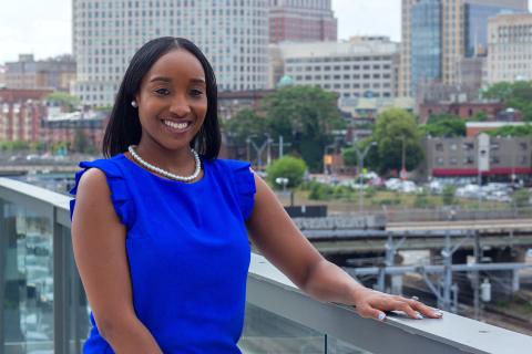 Indira is a Black woman in a bright blue blouse. She poses in front of city buildings.