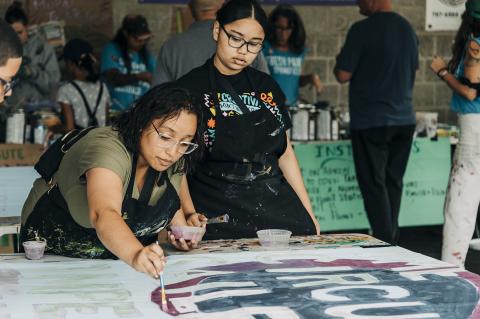 Two light-skinned Black women paint. They wear aprons and more folks are behind them doing the same.