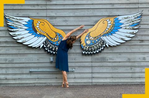 A white woman dances in front of painted wooden wings.