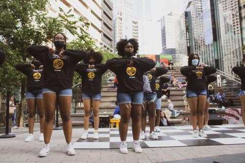 In a city street, a group of Black women, in black and white jersey sweatshirts and denim shorts, dance.