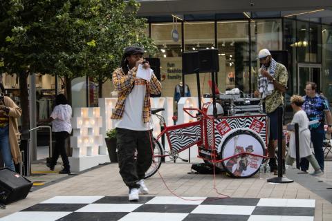 On a black and white checkerboard ground, a man speaks into a mic while another man stands behind a DJ booth on wheels.