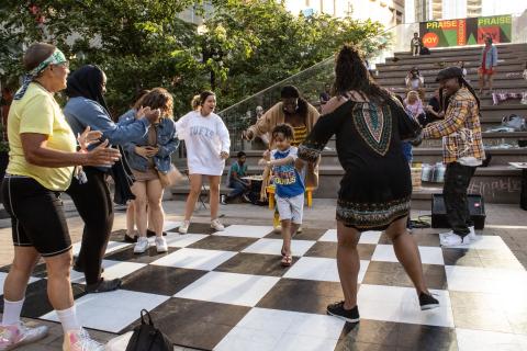 In a circle of folks, a young Black boy dances on top of a black and white checker board ground.