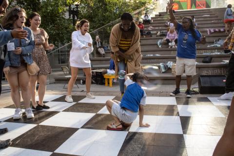 Outside, by stone steps, a circle of folks watch a young, Black girl dance. A blurred bubble floats through the frame.
