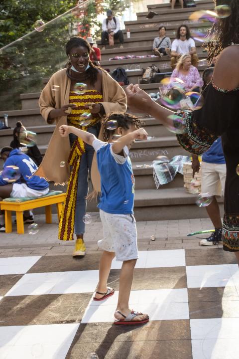 A young person dances in bubbles, while folks look on who are standing nearby or sitting on stone steps.