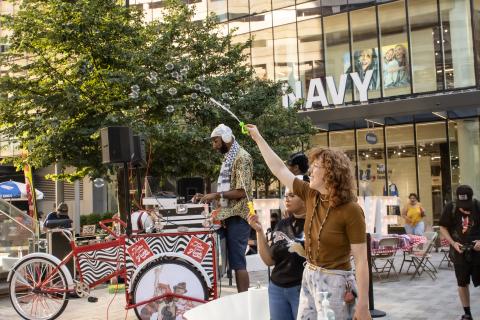 Outside an Old Navy, a white woman, with red hair, holds a bubble wand from which bubbles emit in the breeze.