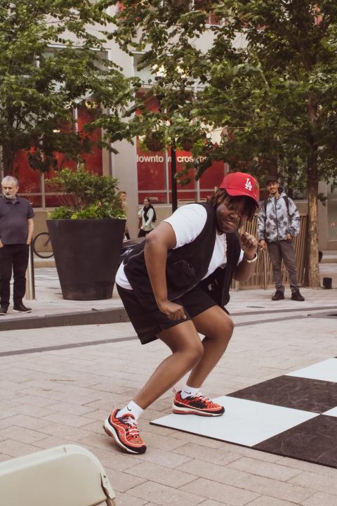On a stone pathway, a young, Black person in a red hat poses, leaning back, with their hand by their mouth.