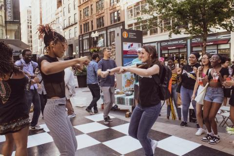 A white woman and a Black woman mirror each other's dance moves, while a crowd looks on.