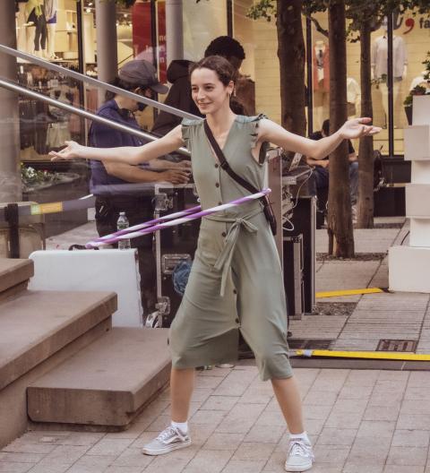 A white woman, in an olive dress, hula hoops.
