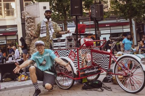 A light-skinned man and a dark-skinned man pose together with a DJ booth on wheels.
