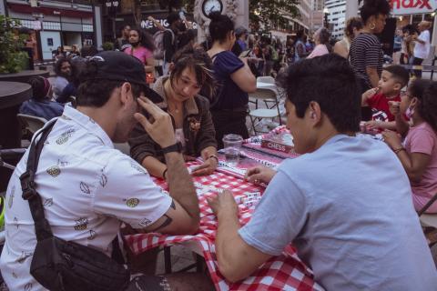 At a picnic table, with red gingham print, three folks play games together.