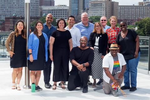 Twelve folks pose on a deck in front of a city skyline.