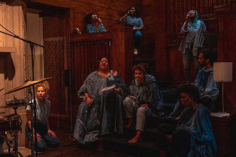 In a wood-panneled home, eight Black folx sing, wearing denim while sitting on some stairs.