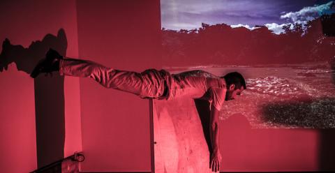 a person balances on their ribcage on a pedestal. A wash of pink light and a video projection of sky