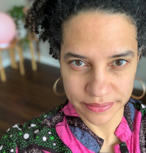 Jess Roseman makes eye contact in front of plants and a globe lamp, in a pink and multicolor patterned top. 