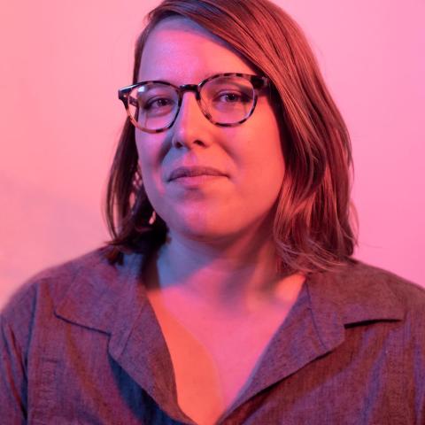 Kelsey poses in a pink light. She has shoulder length hair, is a white lady, and wears glasses.