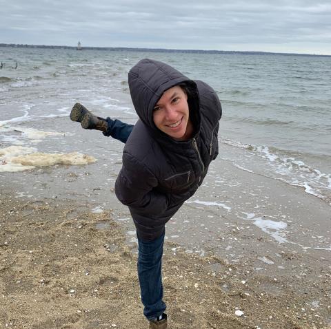 A white woman wearing a hoody standing n arabesque on a beach on an overcast day.