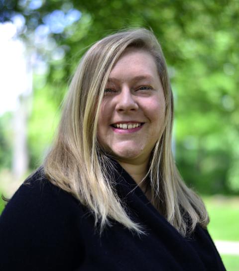 A woman with long straight blonde hair wearing a black shirt; green trees and a blue sky are in the background.