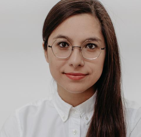 A woman with long straight brown hair over one shoulder wearing round wire glasses and a white button down shirt against a white backdrop.