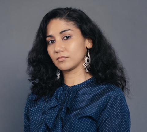 A woman with long dark curly hair wears a blue blouse and dark trousers and sits on a wooden stool with a gray background.