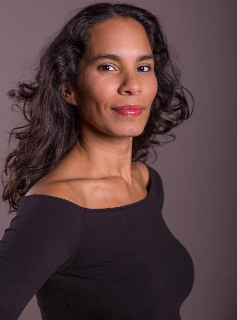 A brown skinned woman with wavy long dark hair wearing a black top with a ballet neckline.