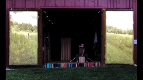 Outside, a man performs in a barn, where the doors have projections of fields on them.