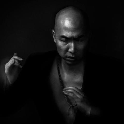 Black and white image of Michael Sakamoto, a Japanese man with a bald head. He poses in a deep v-neck shirt and holds his fists up.