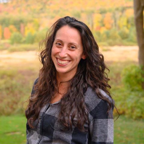 Monica has very long brown hair. She is a white lady. She stands in a field with yellow, red, and green leaved trees behind her.