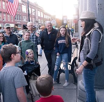 A woman wearing a fedora and a microphone speaks to a small crowd of all ages on street filled with brick buildings.