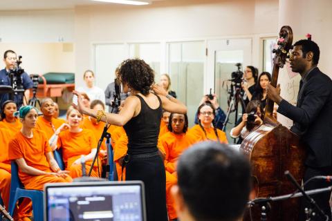 A woman and band with cello conduct a concert in front of a crowd of incarcerated women.