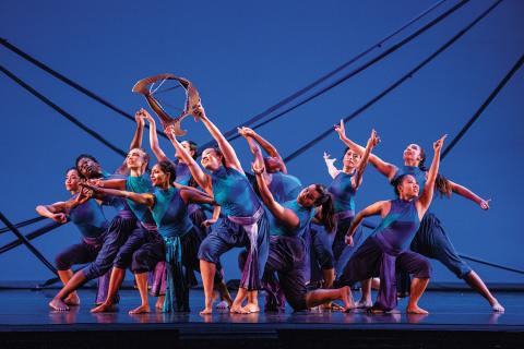 On a stage, a troupe of dancers wear blue and purple in front of a blue backdrop.