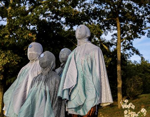 Four people wear bejeweled costumes that cover their faces. They pose plainly in front of trees.