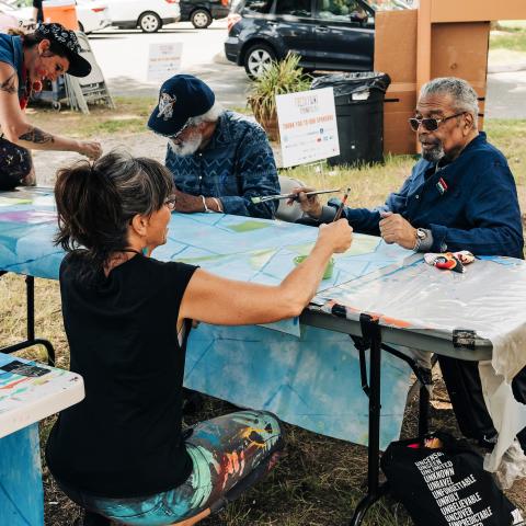Outside, over a folding table, an elderly Black man and a middle-aged white woman paint.