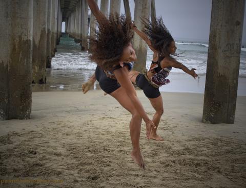 Under a dock, two blacks girls dance in the sand.