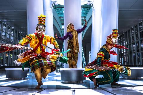 In a mall, with pillars and round, backless benches, a woman poses on a bench and two folks in ornate red, yellow, and green costumes twirl.