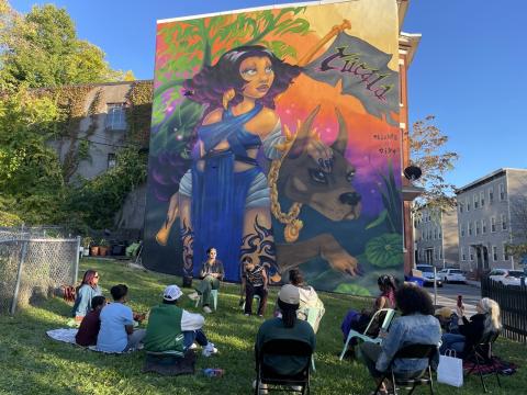 A mural of a woman and a dog with a flag that reads "Cúcala." Below it, a crowd of folks sit in a circle.