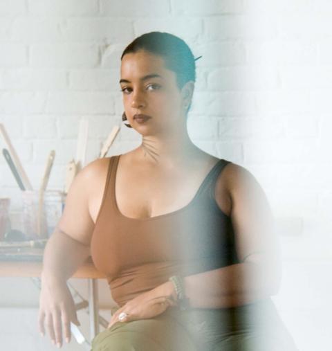 A woman with short dark hear wearing a brown tank top sits in a light room with a plant on a table in the background.