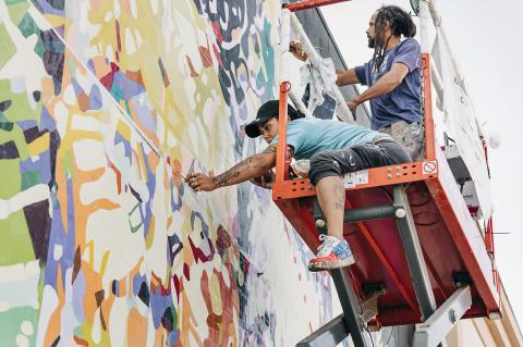 On an orange riser, two folks paint on the side of a building, outside.